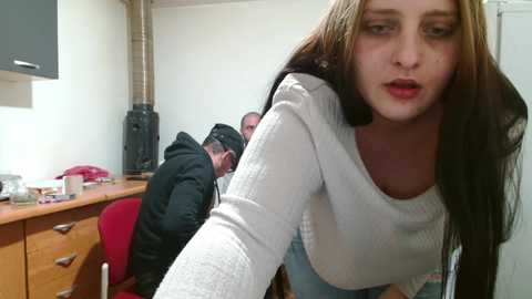 A video shows a young woman with long brown hair, wearing a white sweater, leaning forward in a cluttered room. A man sits behind a desk, partially obscured by a wall-mounted TV.