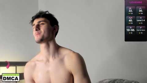 Video of a shirtless, muscular young man with short dark hair, standing in a dimly lit room with a gray wall. A fitness app screen displaying his stats is visible in the top right corner.