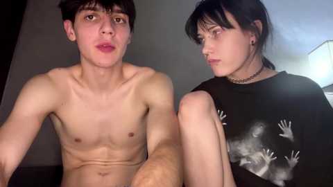 Video of two young people indoors: a shirtless man with short dark hair and a woman with dark hair and a black shirt with hand prints.