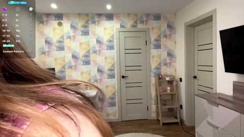 Video of a modern bedroom with a colorful, abstract wallpaper, a woman with long brown hair, a flat-screen TV, and a white door with a handle.