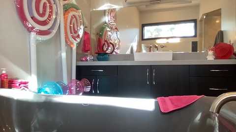 Video of a modern bathroom with a black double sink vanity, white ceramic vessel tub, colorful candy-themed decor, including giant lollipops, and vibrant pink towels.