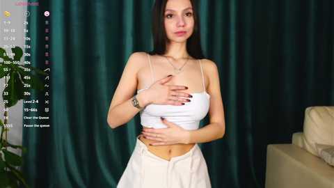 A video of a slender, fair-skinned woman with long black hair, wearing a white spaghetti-strap crop top and white skirt, standing against a dark green curtain backdrop.