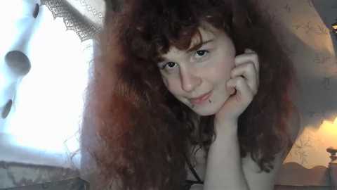 Video of a young woman with long, curly brown hair, light skin, and freckles, lying on a bed with a white lace canopy. She has a subtle smile, looking relaxed.