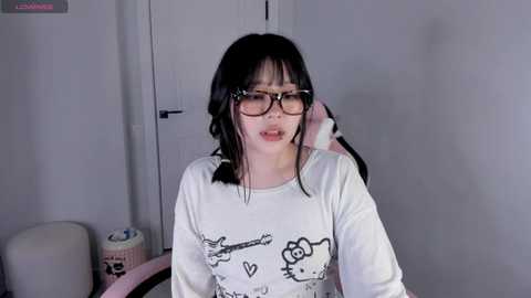 Video of an East Asian woman with long black hair, wearing glasses and a white shirt with cartoon designs, talking on a pink phone in a minimalist room.