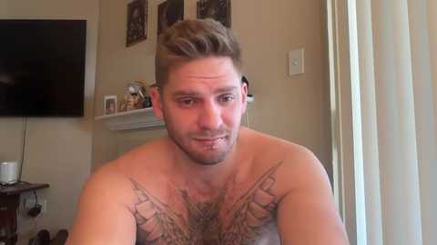 Media: Video of a muscular, light-skinned man with short brown hair, no shirt, showing detailed chest tattoos. Background includes a flat-screen TV, shelves, and framed artwork.