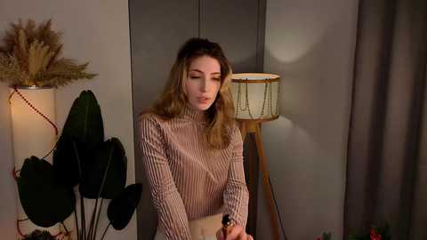 Video of a young woman with wavy blonde hair, wearing a striped sweater, standing in a dimly lit room with a gray wall, potted plant, and a lamp.