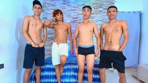 Video of four shirtless, muscular young men with diverse ethnicities, wearing only black briefs, standing in front of a blue patterned cushion and a tiled wall.