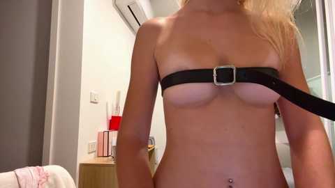 Video of a topless blonde woman in a black leather harness, standing in a modern, minimalistic bedroom with white walls, an air conditioner, and a wooden dresser.