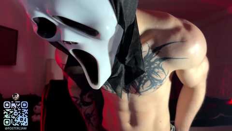 Video of a shirtless, muscular man with a large black tribal tattoo on his chest, wearing a white, mask-like face covering, in a dimly lit room with red lighting.