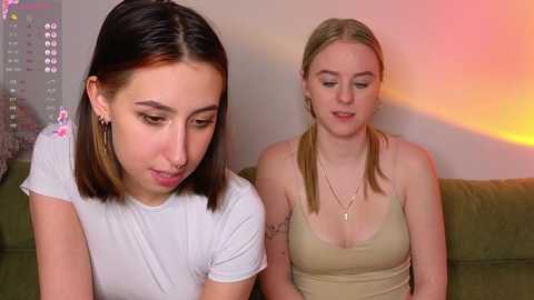 Video of two young women with straight hair, one with a nose ring, wearing white and beige tank tops, sitting on a green couch, against a colorful wall.