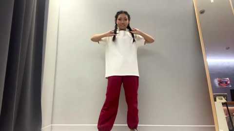 Video of an East Asian woman with long black hair in braids, wearing a white t-shirt and red pants, standing against a gray wall in a well-lit room.