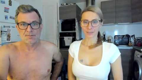 Video of a middle-aged, fair-skinned couple with glasses; man shirtless, woman in white top, both smiling in a modern kitchen with stainless steel appliances.