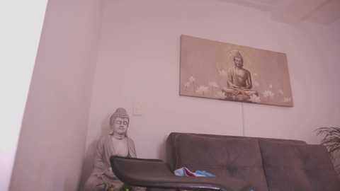 A video of a minimalist room with a serene, beige Buddha statue on a pedestal, a large, framed Buddha painting on a white wall, and a dark brown couch with a blue and white pillow.