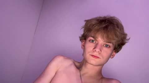 Video of a young, fair-skinned person with short, tousled brown hair, green eyes, and a slight smile. They are topless, standing against a plain, light purple wall.