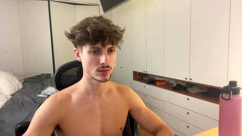 Video of a shirtless young man with curly dark hair, sitting in a chair, in a minimalist bedroom with white wardrobe, bed, and mirror.