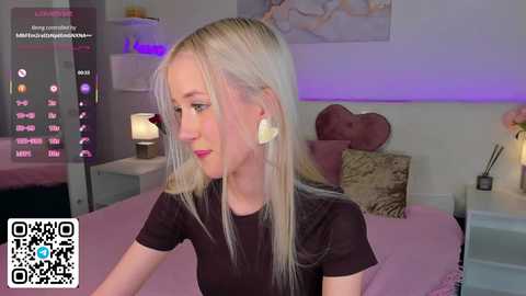 Video of a blonde woman with fair skin wearing a black top and large white earrings, sitting on a bed in a modern, dimly lit bedroom.