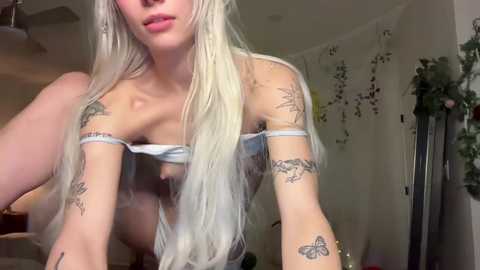 Video of a young, light-skinned woman with platinum blonde hair, wearing a sheer, light blue top revealing her small breasts and multiple tattoos on her arms. The background shows a cozy, dimly-lit room with hanging plants and a mirror.