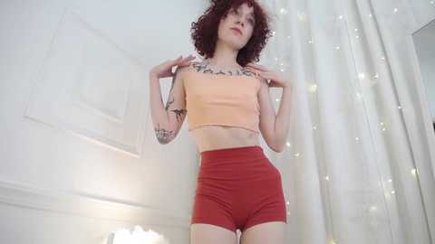 Video of a slender, pale-skinned woman with curly red hair wearing a beige crop top and red shorts. She stands confidently indoors, adorned with tattoos, against a white wall and fairy lights.