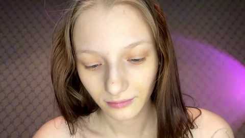 Video of a young, light-skinned girl with long, wet, brown hair, wearing no top, sitting against a textured, dark background with a purple light illuminating her face.