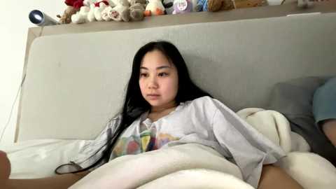 Video of a young Asian woman with long black hair, wearing a white shirt, lying in bed with a beige blanket. Toys and stuffed animals are on the bed's headboard.