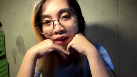 Video of a young Asian woman with straight, light brown hair, wearing glasses, a light blue top, and resting her chin on her hands. Background shows a plain, beige wall with green blinds partially visible.