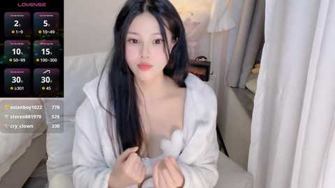 A video of an Asian woman with long black hair, wearing a white robe, sitting on a bed with white sheets. The background includes a window with white curtains and a digital screen displaying weather and social media information.