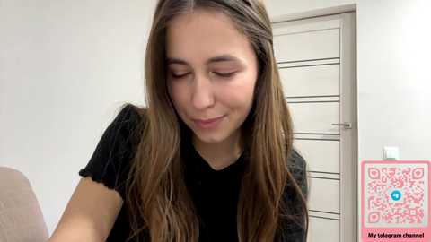 Media: A video of a young woman with long, straight brown hair and fair skin, wearing a black top, smiling softly. The background features a white wall and a closed door.