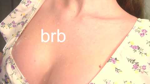 A close-up video of a woman's bare chest with fair skin, showing a small mole and a tattoo on her left breast. She wears a light-colored floral top, slightly open, revealing cleavage.