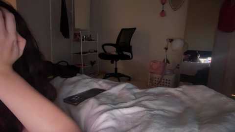 Video of a dimly-lit bedroom with a person in bed, covered by a white blanket, and a black chair nearby; the room has minimalistic decor.