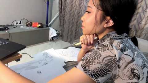 Video of an East Asian woman with black hair, dressed in a patterned shirt, sitting at a cluttered desk with papers and a laptop.