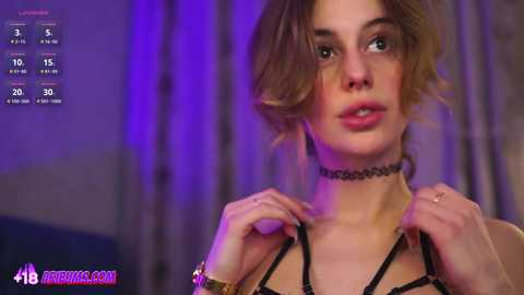 Video of a young woman with short, tousled brown hair, wearing a black lace choker and a strappy black top, with purple lighting in the background.