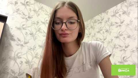 Video of a young woman with straight brown hair, wearing glasses, white t-shirt, and floral wallpaper background.