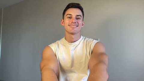 Video of a young, muscular man with short brown hair, wearing a white sleeveless shirt, smiling against a plain, light gray wall.