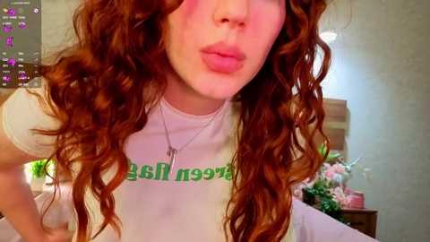 Media: Video of a fair-skinned woman with long, curly red hair, wearing a white t-shirt with green text, puckering her lips, in a cozy, dimly-lit room with floral arrangements.