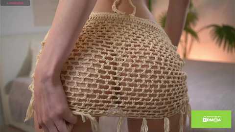 A video of a woman's lower back and buttocks, wearing a beige, crocheted skirt with frayed edges, standing indoors with blurred background. The image includes a green text box with \"\u0411\u0435\u043b\u0430\u044f \u0414\u043e\u043c\u043d\u0430\" and a website URL.
