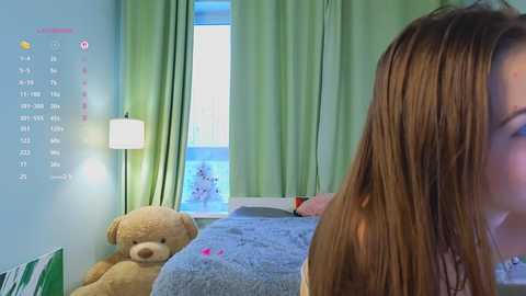 Video of a young woman with long brown hair, partially visible, in a cozy bedroom with green walls, a teddy bear, and a calendar on the left.