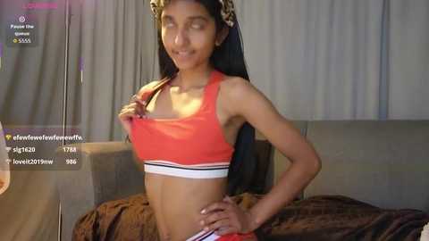 A video of a slender South Asian woman with long black hair, wearing an orange sports bra and striped shorts, standing confidently with her hand on her hip in a dimly lit bedroom.