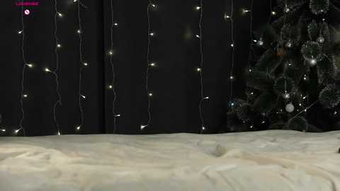 A video of a Christmas scene: a dark curtain with white string lights, a snow-covered Christmas tree, and a white bedspread.