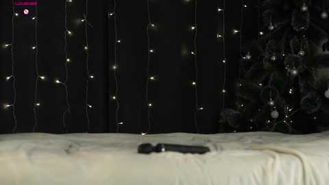 A video shows a bed with a beige sheet and a black remote control placed on it. The background features a dark curtain with twinkling white fairy lights and a partially visible Christmas tree adorned with white ornaments.