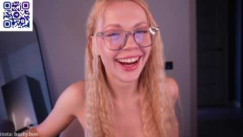 Video of a blonde, fair-skinned woman with glasses, smiling widely, wearing a strapless top. Background shows a modern, dimly lit room with a QR code watermark.