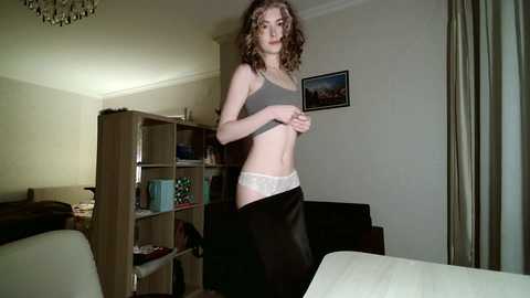 Video of a slim, light-skinned woman with curly brown hair, wearing a grey sports bra and black skirt, standing in a dimly lit living room with beige walls, wooden bookshelves, and a framed landscape painting.