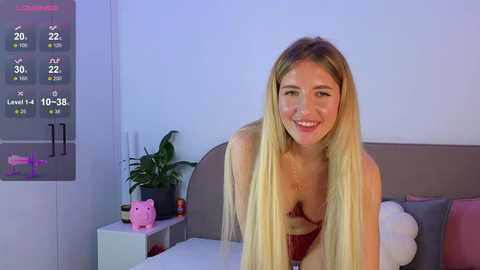 Video of a smiling blonde woman in a red bra, standing in a modern bedroom with a digital clock, plant, and pink cat figurine on a white nightstand.