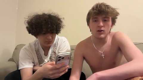 A video of two young men, one with curly hair, the other shirtless, sitting on a couch, focusing on a smartphone.