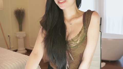 Video of a smiling woman with long, straight black hair, wearing a sleeveless olive green lace top, sitting in a softly lit, minimalist bedroom with a white bed and a vase of decorative grasses.