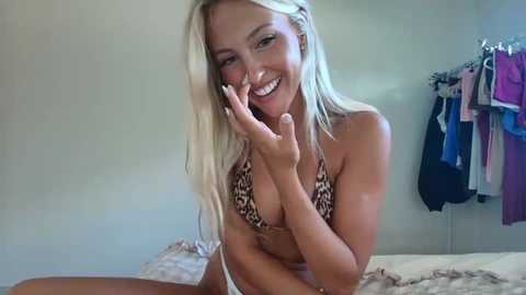 Video of a smiling, slender, blonde woman in a leopard-print bikini, with a hint of tan, posing on a bed in a brightly lit room with hanging clothes.