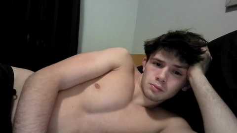 Video of a young, shirtless man with fair skin and dark hair, lying on a bed with black sheets, looking tired and resting his head on his hand.
