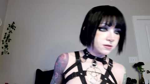 Video of a pale-skinned woman with black bob haircut, wearing black leather harness and choker, tattoos on her arm, in a dimly lit room with white walls and black decor.