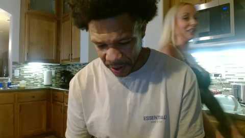 Video of a man with a large afro, wearing a white Essential Elements T-shirt, smiling in a modern kitchen with wooden cabinets, a microwave, and a blonde woman in the background.