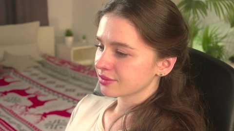 Video of a young woman with long brown hair, fair skin, and subtle makeup, sitting in a chair in a cozy, sunlit room with a bed adorned with red and white patterned bedding.
