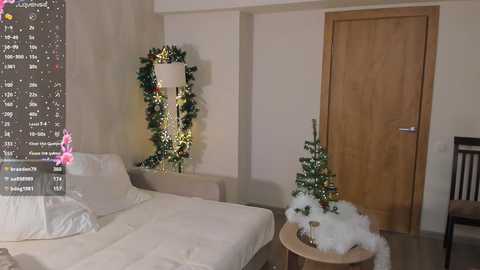 Video of a minimalist, beige-walled bedroom with a white bed, a wooden door, a Christmas tree, a white lamp, and a pink heart symbol on a calendar.